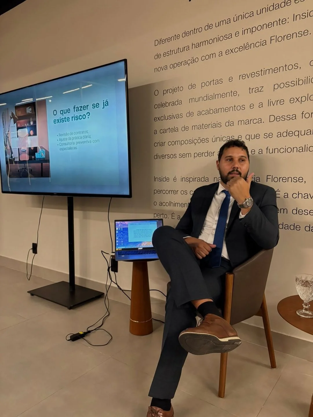 Advogado Luís Fernando, do escritório P. Lasmar Advocacia, sentado ao lado de um telão com apresentação sobre prevenção de riscos trabalhistas.
